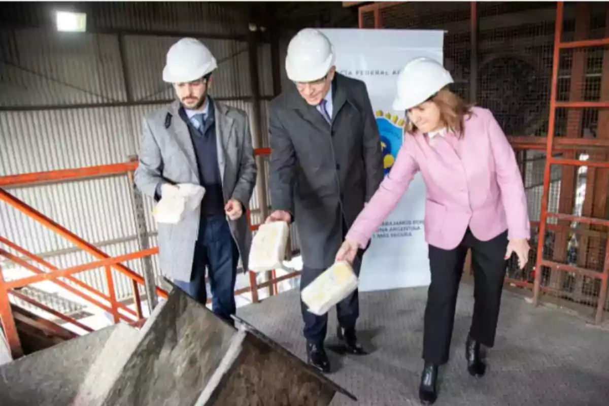 Tres personas con casco y ropa formal depositan paquetes en una tolva industrial dentro de una fábrica