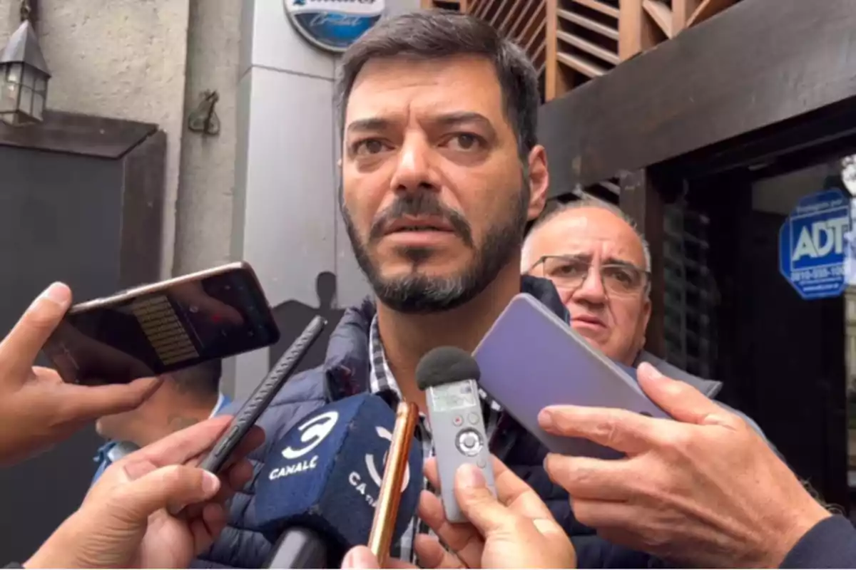 A man with a beard and short hair is interviewed by several journalists holding microphones and phones in front of him while another man watches in the background. A man with a beard and short hair is interviewed by several journalists holding microphones and phones in front of him while another man watches in the background.