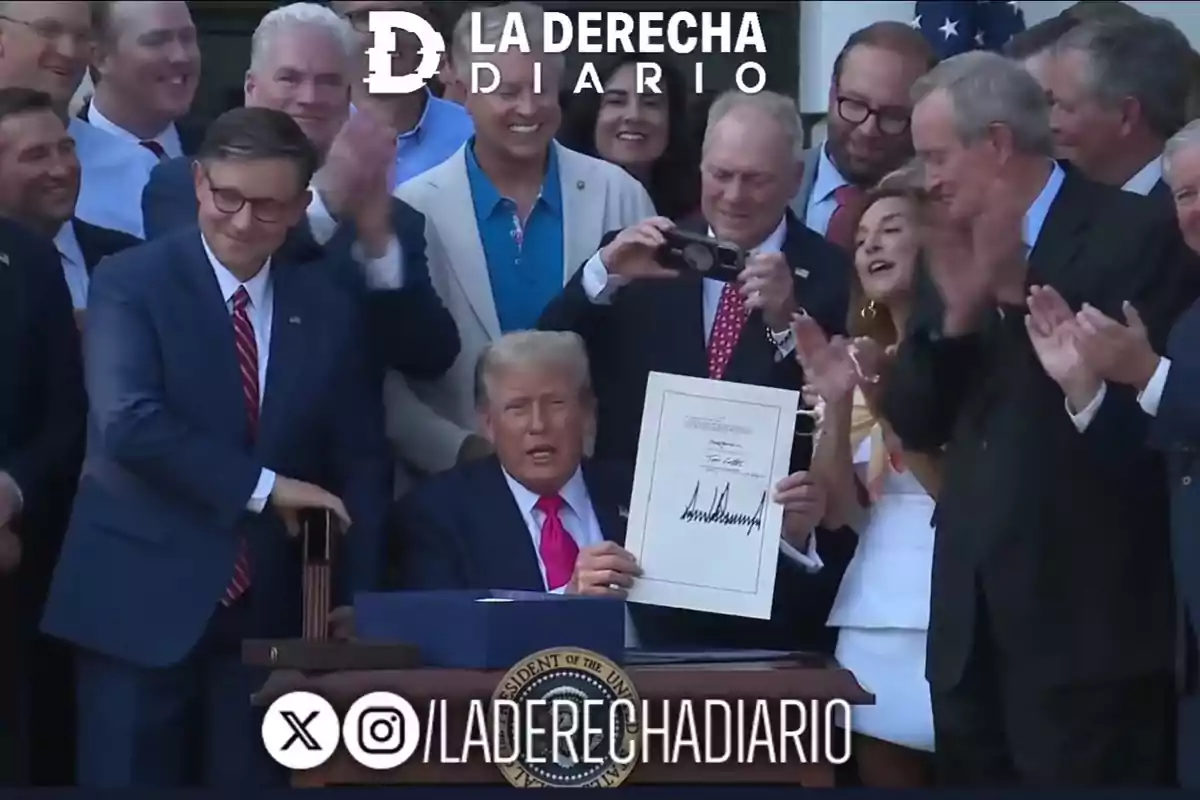 A group of people applauds while a man in the center holds a signed document and sits in front of a desk with the presidential seal of the United States. A group of people applauds while a man in the center holds a signed document and sits in front of a desk with the presidential seal of the United States.