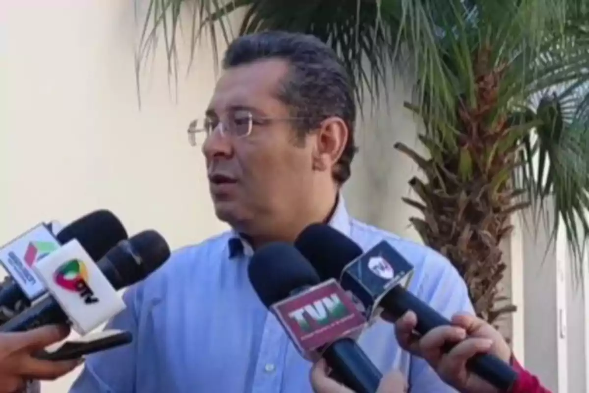 Man with glasses and a light blue jersey giving an interview to several media outlets outdoors with a palm tree in the background Man with glasses and a light blue jersey giving an interview to several media outlets outdoors with a palm tree in the background