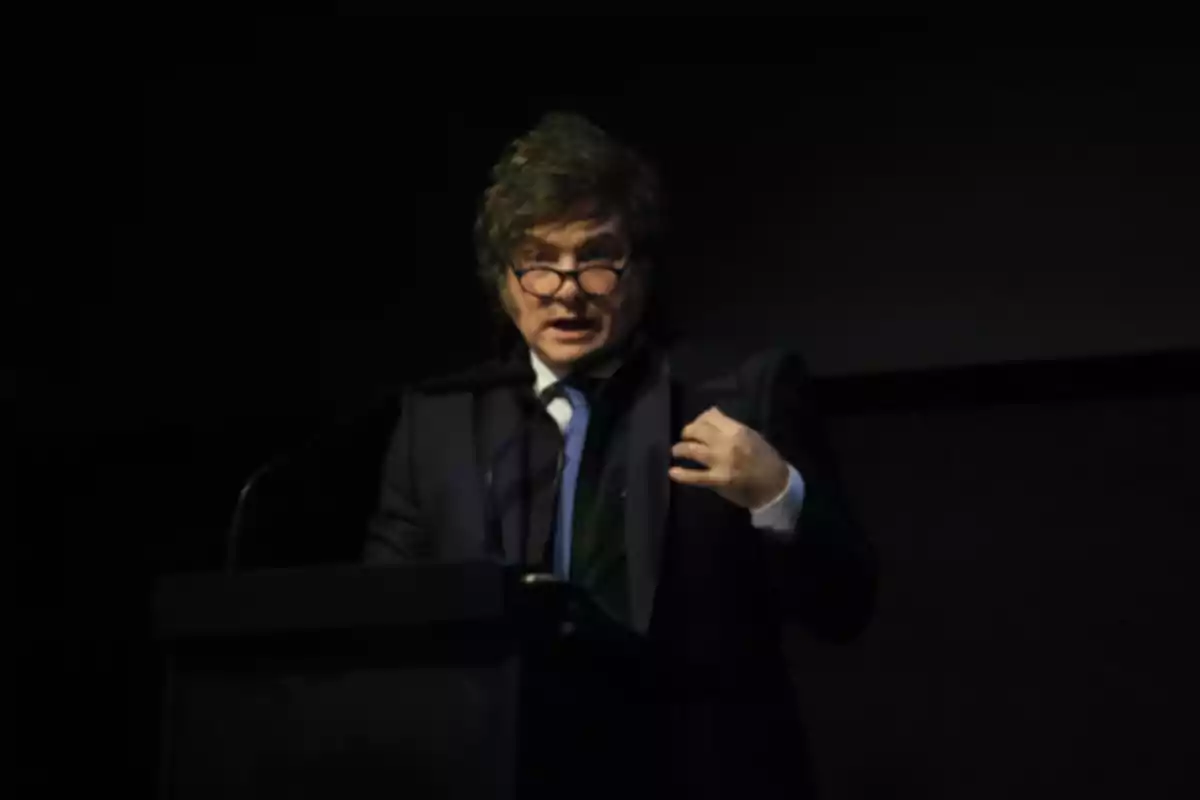 Hombre con gafas y traje oscuro hablando en un podio en un ambiente con poca luz