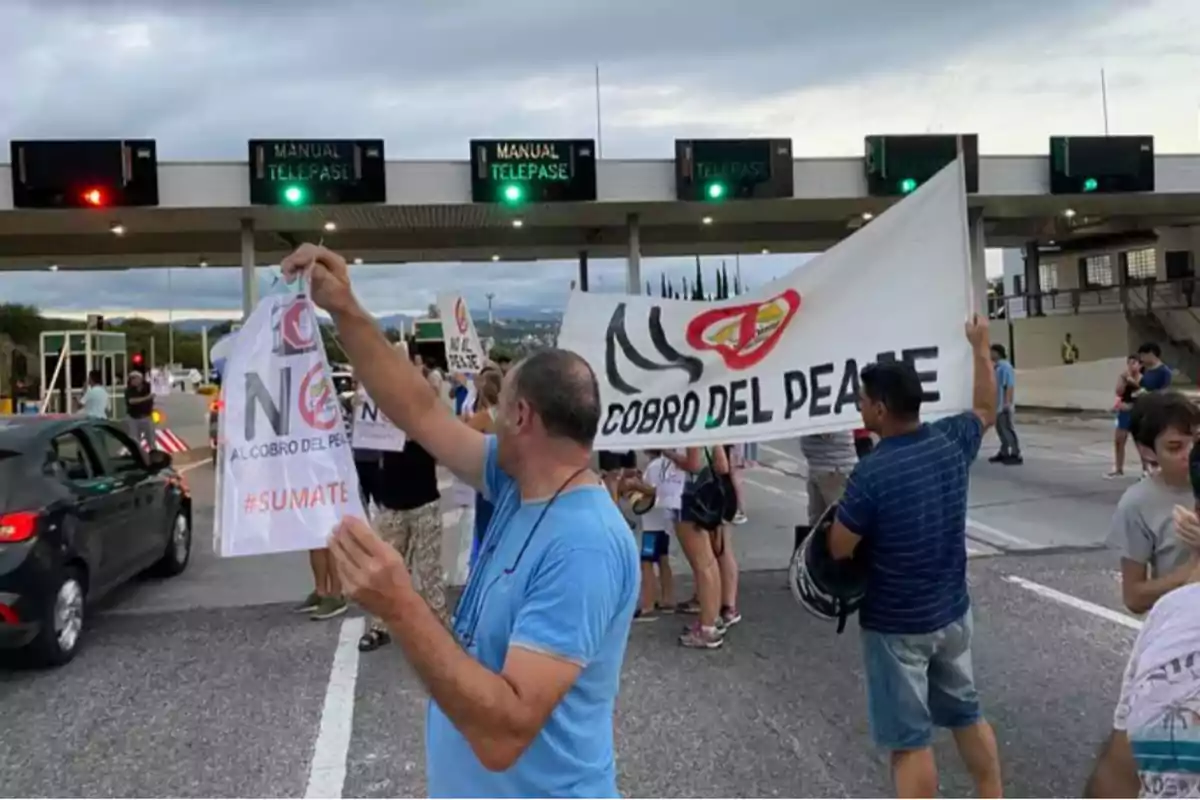 Personas protestando en una cabina de peaje con carteles y una pancarta que dice no al cobro del peaje mientras algunos autos pasan por el lugar