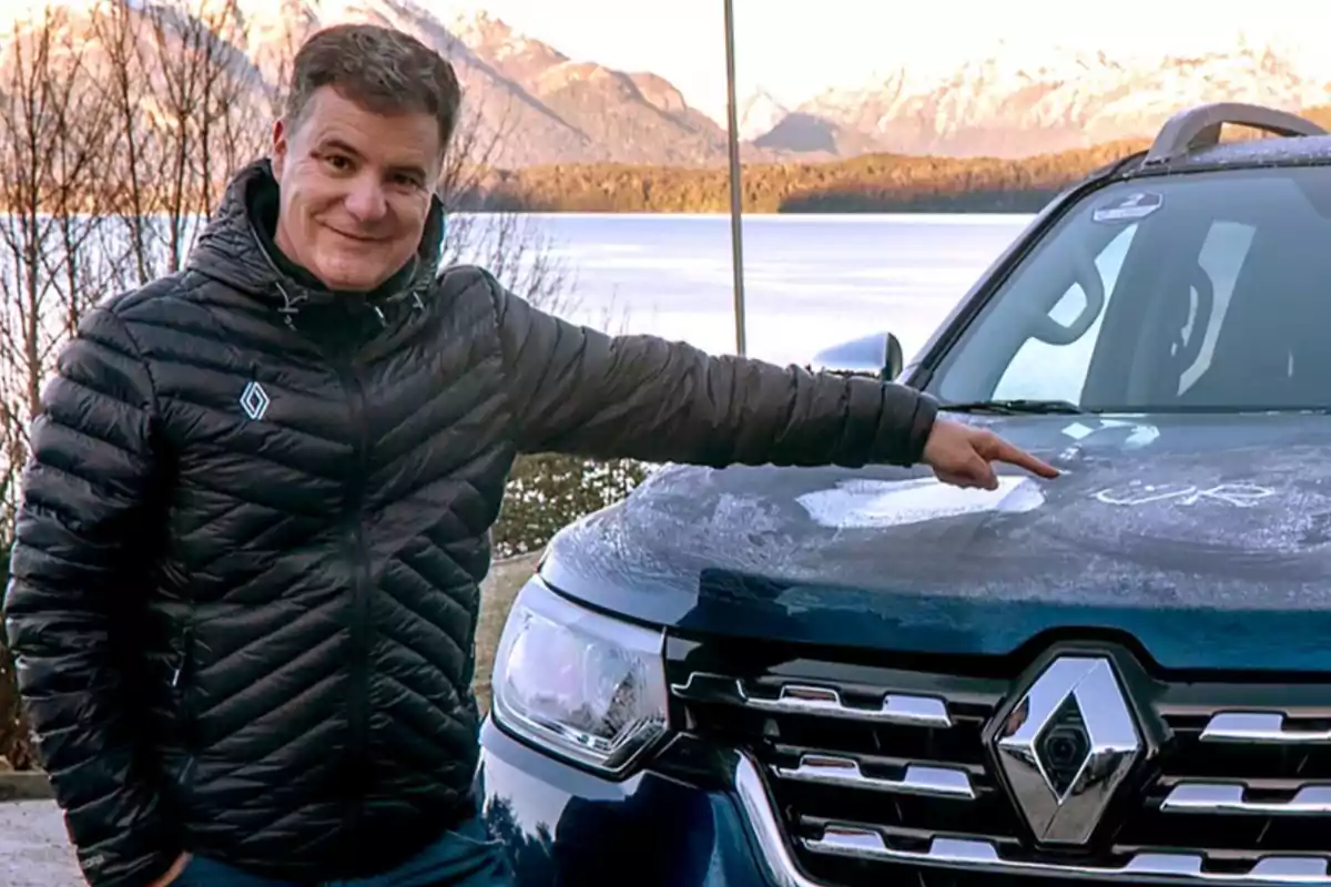 Smiling man in a black jacket pointing at the hood of a blue Renault pickup truck with snow and mountains in the background