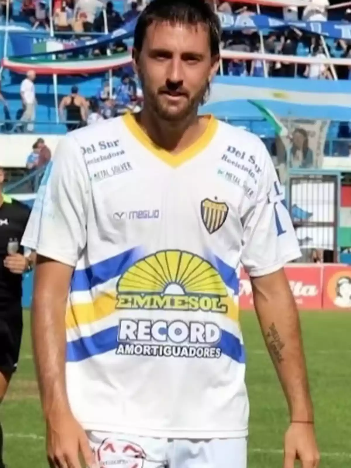Soccer player wearing a white jersey from a team with yellow and blue details posing on a field with spectators in the stands in the background Soccer player wearing a white jersey from a team with yellow and blue details posing on a field with spectators in the stands in the background