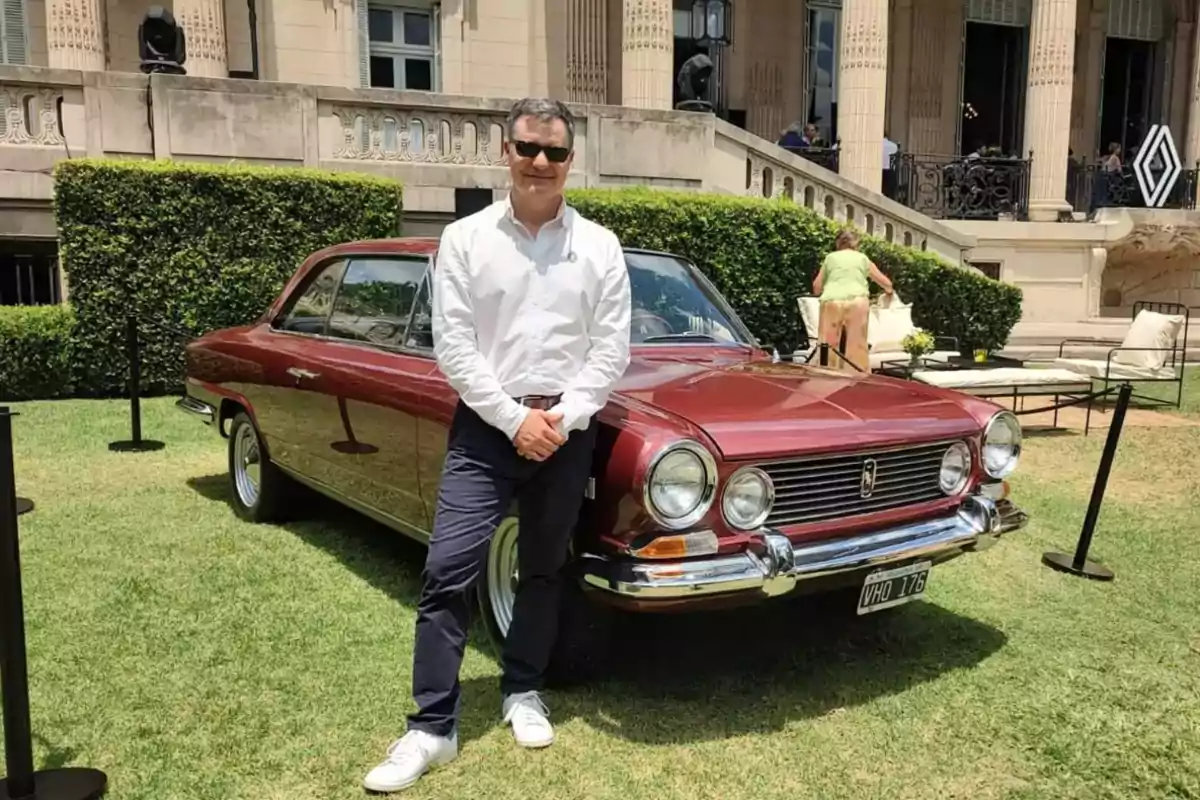 Man wearing a white jersey and sunglasses posing in front of a burgundy classic car in a garden with an elegant building in the background Man wearing a white jersey and sunglasses posing in front of a burgundy classic car in a garden with an elegant building in the background
