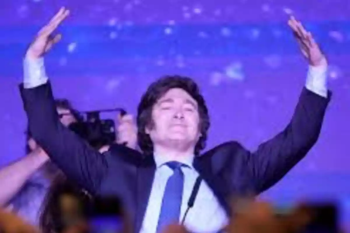 Man in a suit and tie raising both arms on a stage with a purple background Man in a suit and tie raising both arms on a stage with a purple background