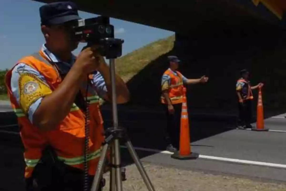Traffic police officers wearing orange vests are conducting a speed check on a highway using a radar and orange cones.