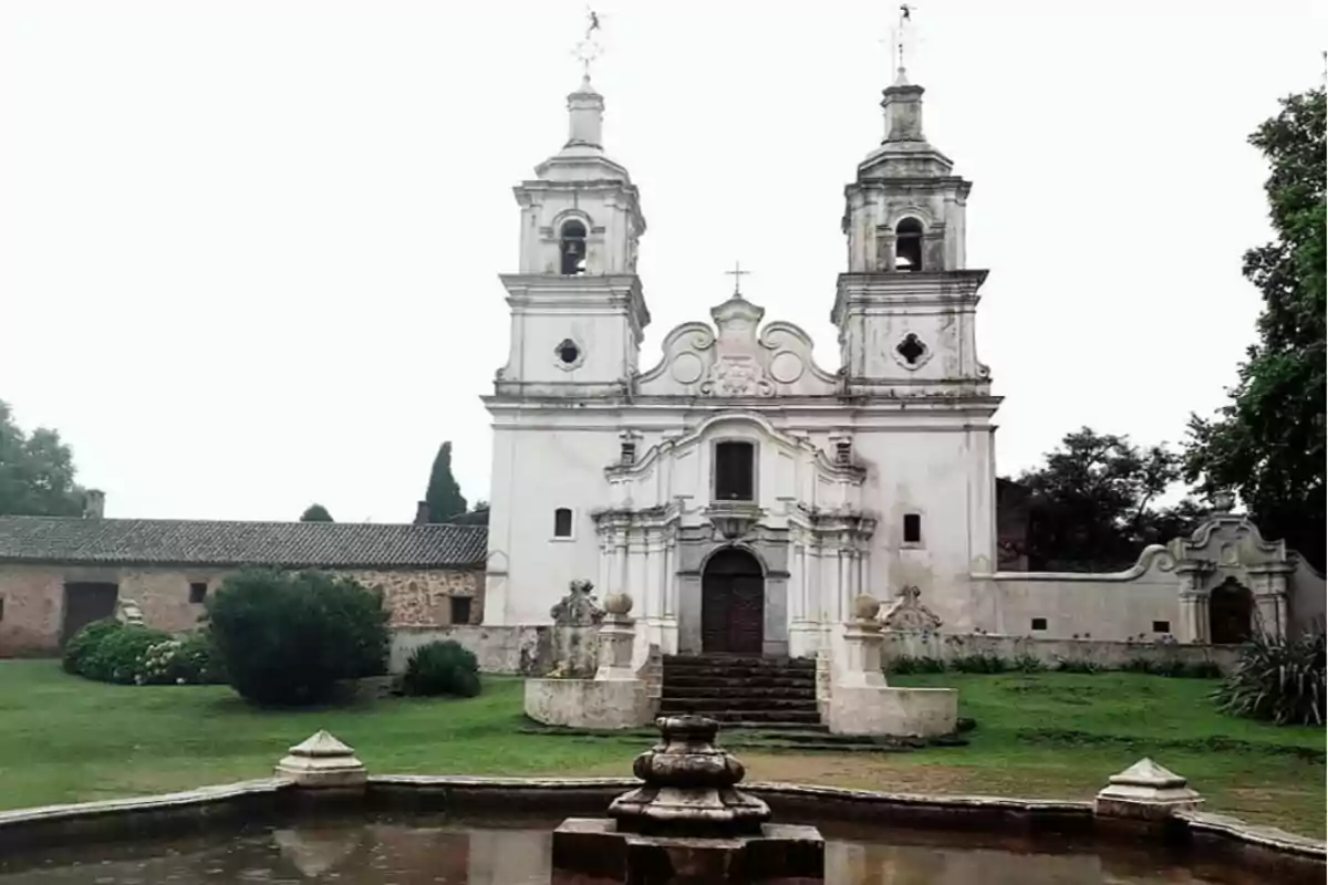 Facade of an old colonial-style church with two towers and a fountain in the foreground surrounded by grass and vegetation Facade of an old colonial-style church with two towers and a fountain in the foreground surrounded by grass and vegetation