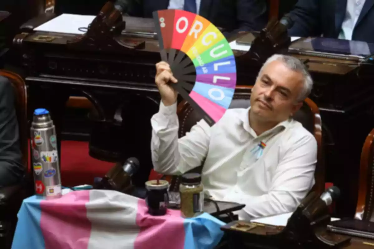 Persona sentada en un recinto parlamentario sosteniendo un abanico de colores con la palabra orgullo mientras en el escritorio hay una bandera trans y una botella con calcomanías de la diversidad