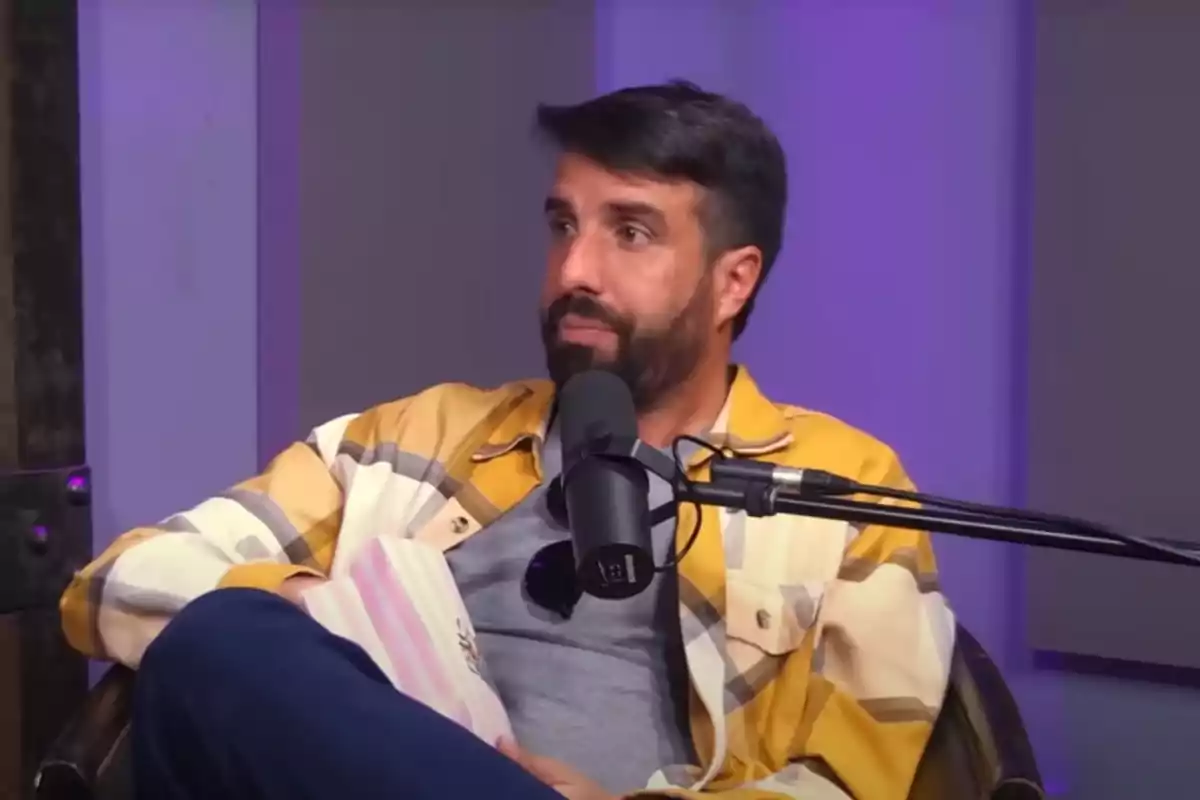 Hombre con barba y cabello oscuro sentado en un estudio de grabación con micrófono frente a él, usando camisa de cuadros amarillos y fondo iluminado en tonos morados Hombre con barba y cabello oscuro sentado en un estudio de grabación con micrófono frente a él, usando camisa de cuadros amarillos y fondo iluminado en tonos morados
