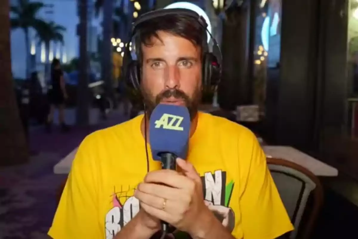 Hombre con barba y auriculares sosteniendo un micrófono azul con letras amarillas, viste una camiseta amarilla y está sentado en una terraza al aire libre por la noche Hombre con barba y auriculares sosteniendo un micrófono azul con letras amarillas, viste una camiseta amarilla y está sentado en una terraza al aire libre por la noche