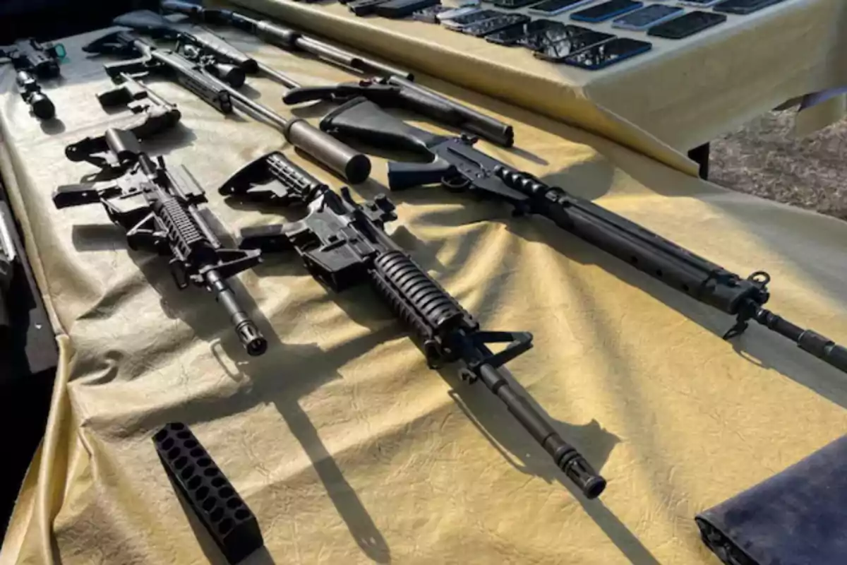 Several long guns and handguns, along with mobile phones, are arranged on a table covered with a yellow tablecloth. Several long guns and handguns, along with mobile phones, are arranged on a table covered with a yellow tablecloth.