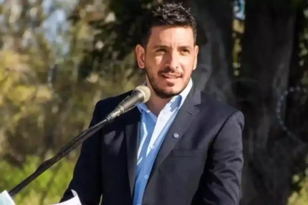 Hombre de cabello corto y barba usando saco azul y camisa clara hablando frente a un micrófono al aire libre