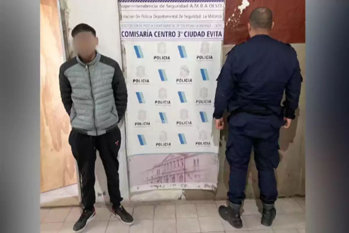 Un joven con el rostro difuminado está de pie junto a un policía de espaldas frente a un cartel de la comisaría centro 3 de Ciudad Evita Un joven con el rostro difuminado está de pie junto a un policía de espaldas frente a un cartel de la comisaría centro 3 de Ciudad Evita