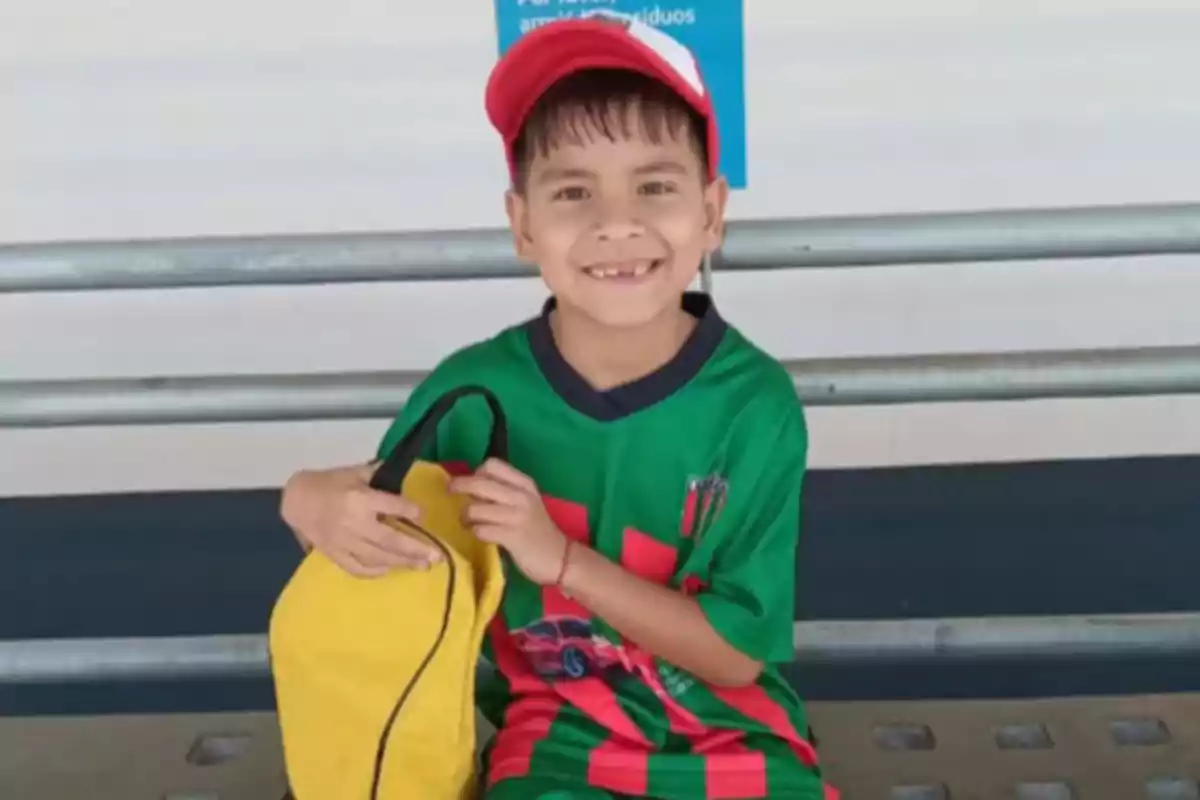 Smiling boy with a red cap and green jersey sitting on a bench holding a yellow backpack