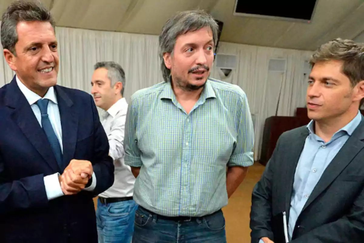 Tres hombres conversan en un ambiente interior con cortinas blancas de fondo, dos de ellos visten traje y el del centro lleva una camisa a cuadros.