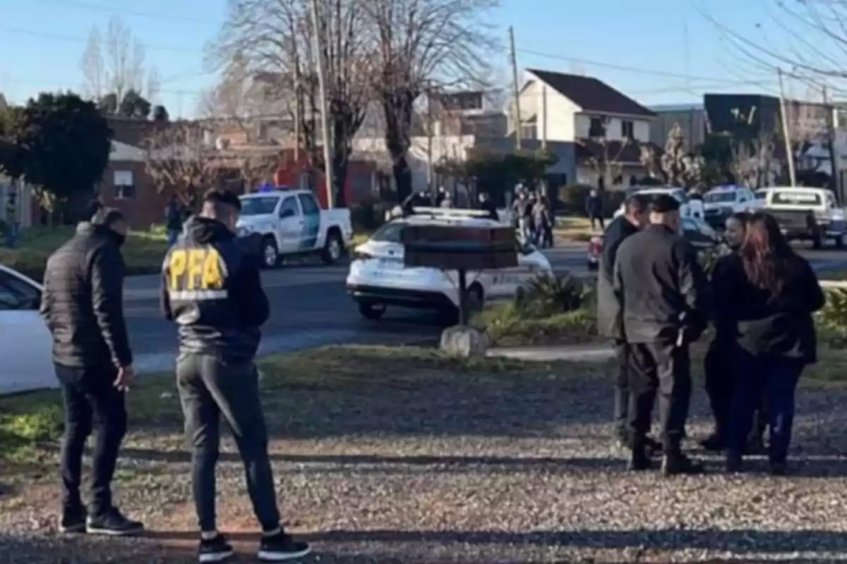 Varios agentes policiales conversan en una calle de barrio residencial con patrulleros y camionetas estacionados al fondo en un día soleado.