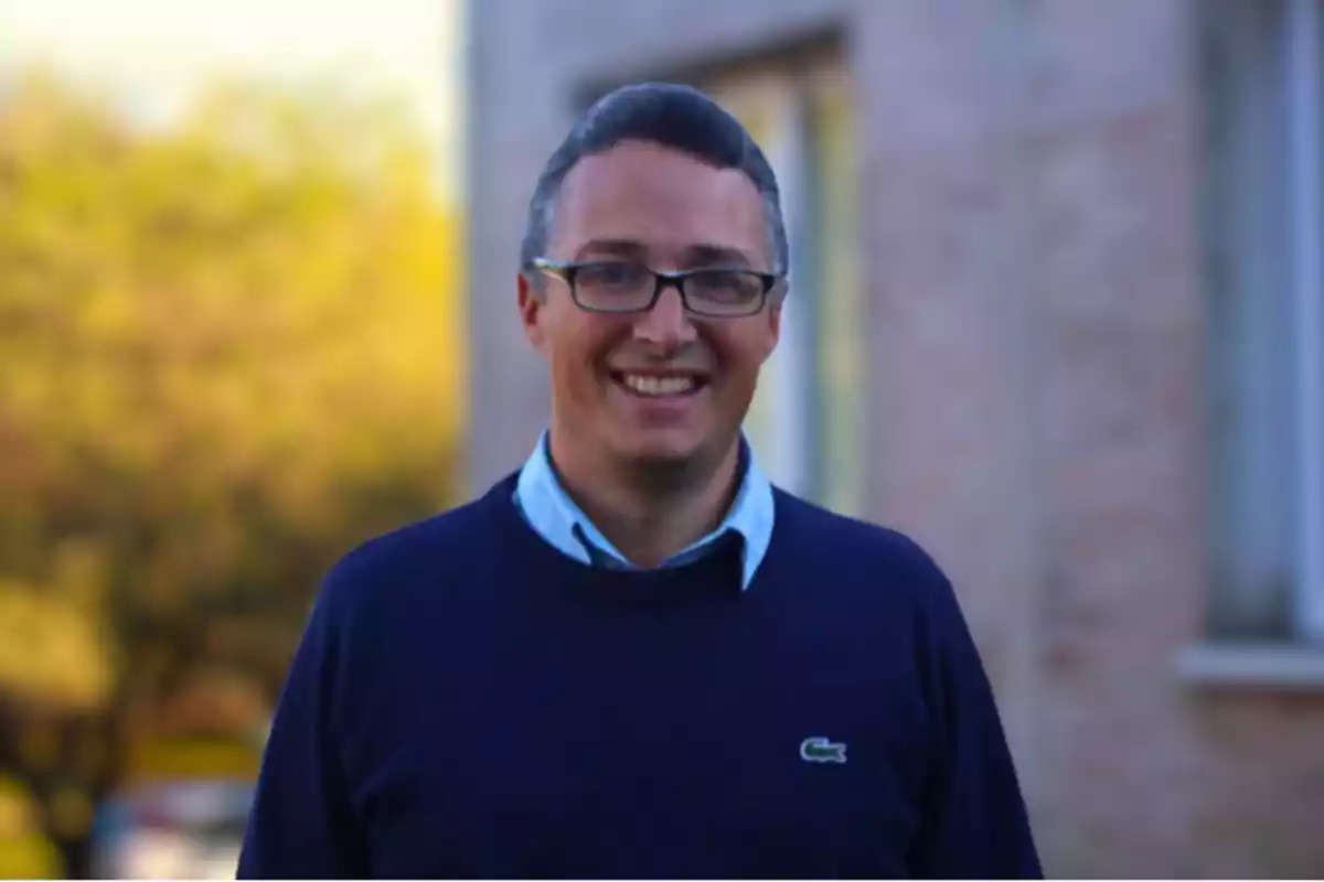 Hombre con gafas y suéter azul sonriendo al aire libre frente a un edificio desenfocado Hombre con gafas y suéter azul sonriendo al aire libre frente a un edificio desenfocado
