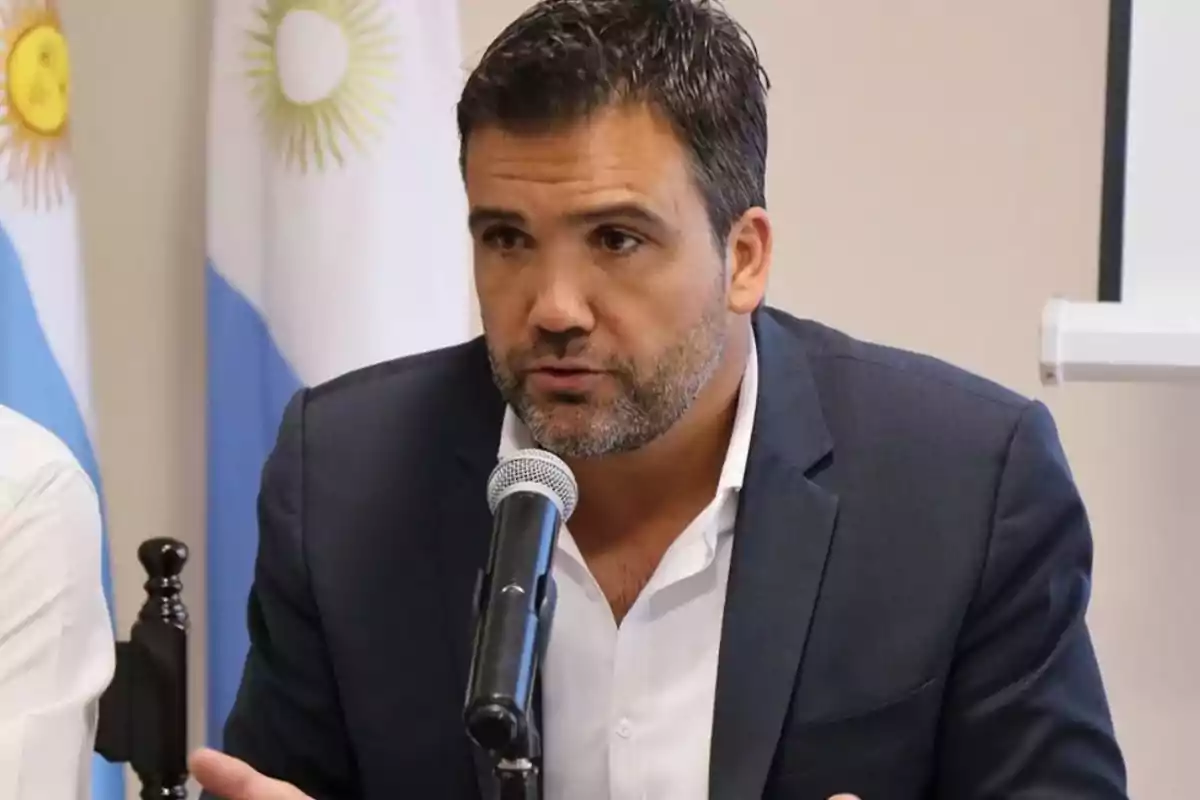 Hombre de cabello corto y barba hablando frente a un micrófono con banderas de Argentina de fondo