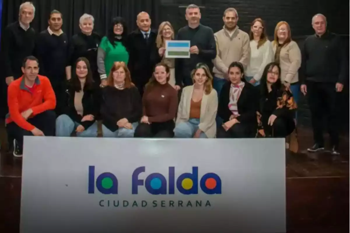 Group of people posing behind a sign that says "La Falda ciudad serrana" in an indoor setting