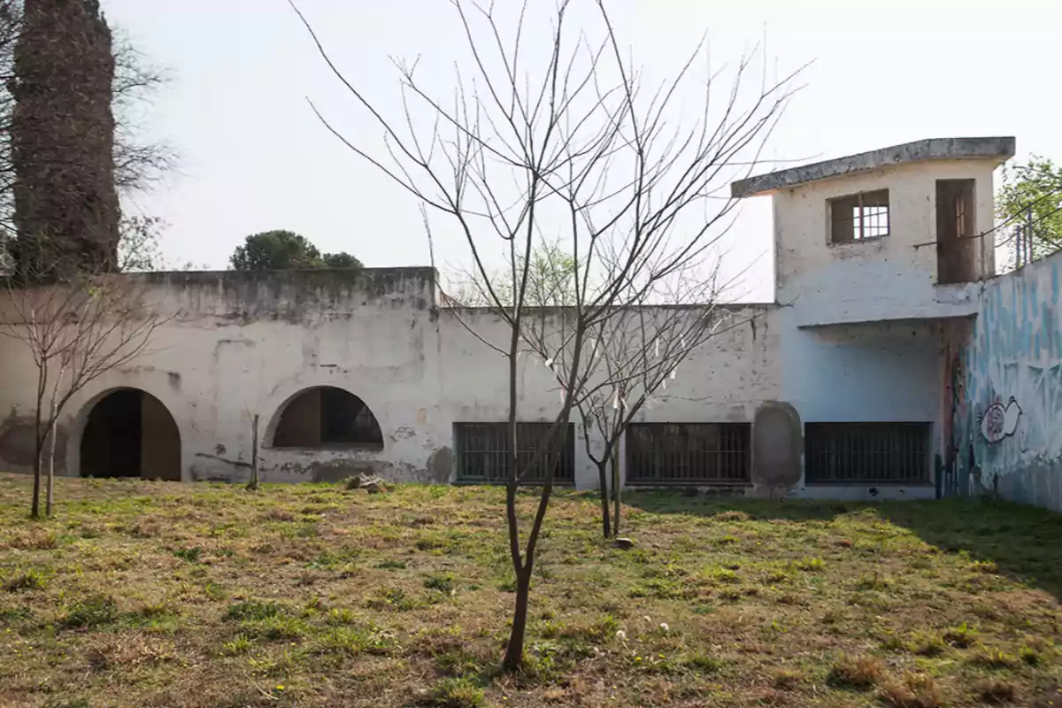 Edificio antiguo y deteriorado con paredes blancas, ventanas enrejadas y árboles sin hojas en un patio de césped seco Edificio antiguo y deteriorado con paredes blancas, ventanas enrejadas y árboles sin hojas en un patio de césped seco