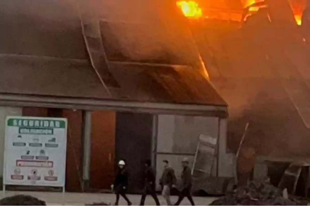 Un grupo de personas con cascos camina frente a un edificio industrial en llamas mientras el fuego y el humo salen del techo Un grupo de personas con cascos camina frente a un edificio industrial en llamas mientras el fuego y el humo salen del techo