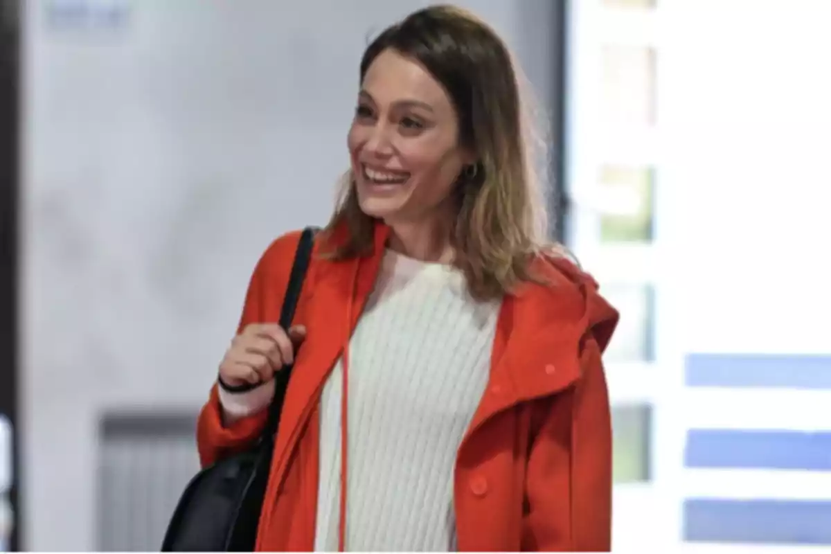 Young woman smiling, wearing an orange coat and a black bag over her shoulder, dressed in a white sweater with her hair down. Young woman smiling, wearing an orange coat and a black bag over her shoulder, dressed in a white sweater with her hair down.