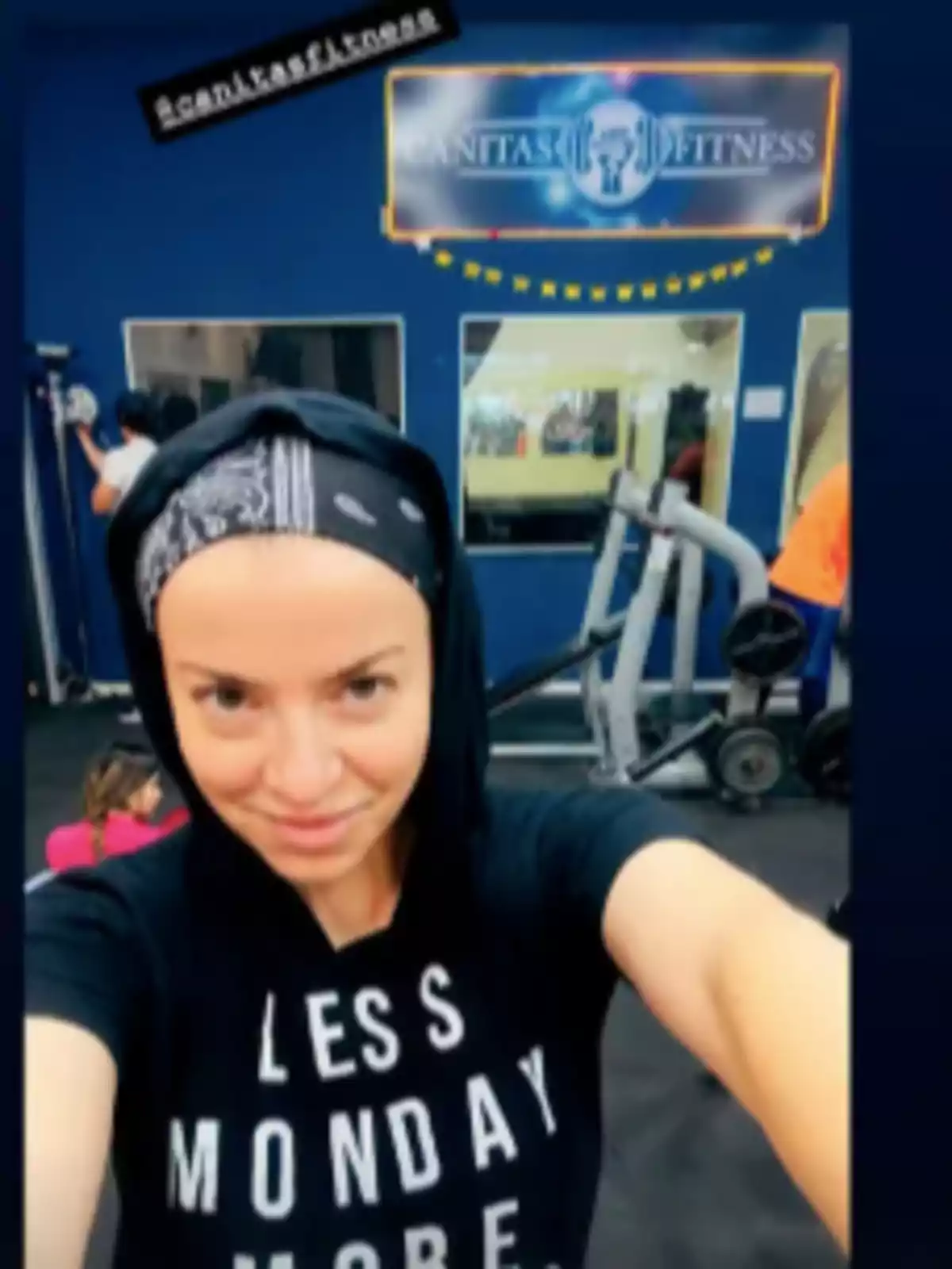 Mujer tomándose una selfie en un gimnasio con una camiseta negra que tiene letras blancas y una banda en la cabeza, al fondo se ven máquinas de ejercicio y un letrero que dice Sanitas Fitness Mujer tomándose una selfie en un gimnasio con una camiseta negra que tiene letras blancas y una banda en la cabeza, al fondo se ven máquinas de ejercicio y un letrero que dice Sanitas Fitness