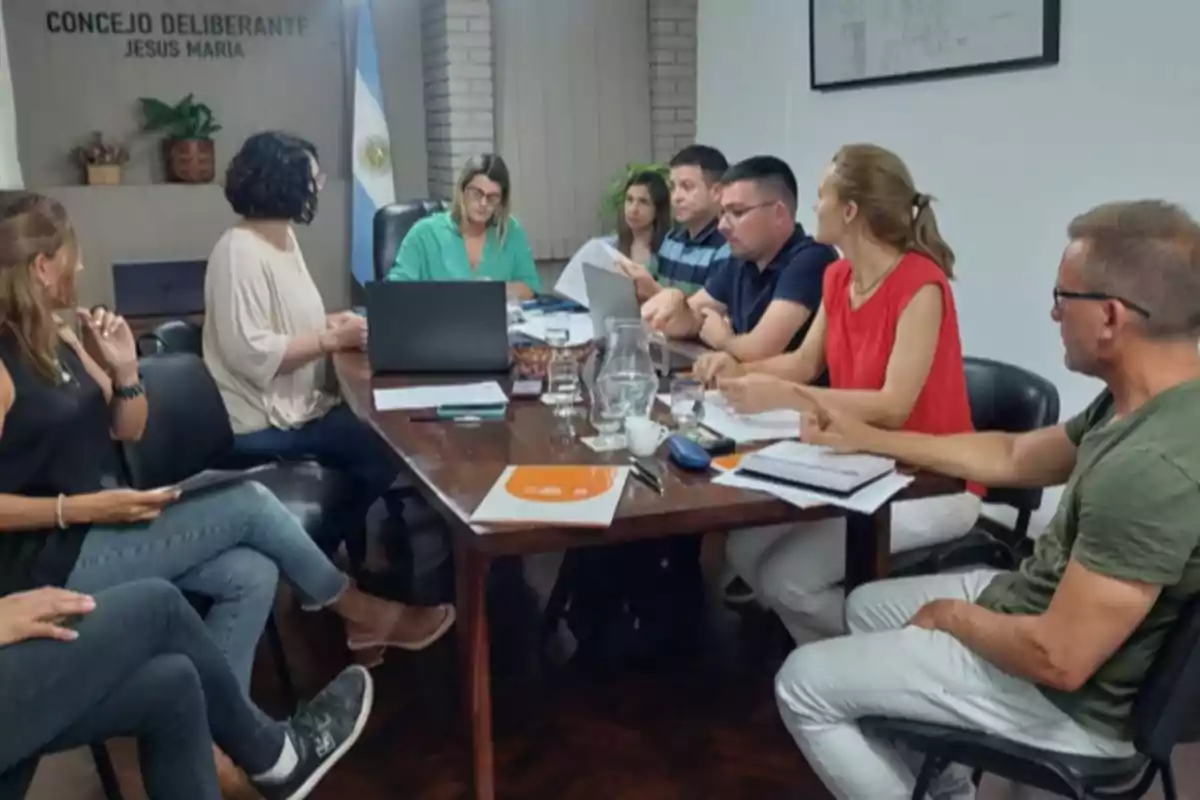 Varias personas sentadas alrededor de una mesa en una sala de reuniones con papeles, computadoras y vasos de agua, participando en una discusión grupal en el Concejo Deliberante de Jesús María.