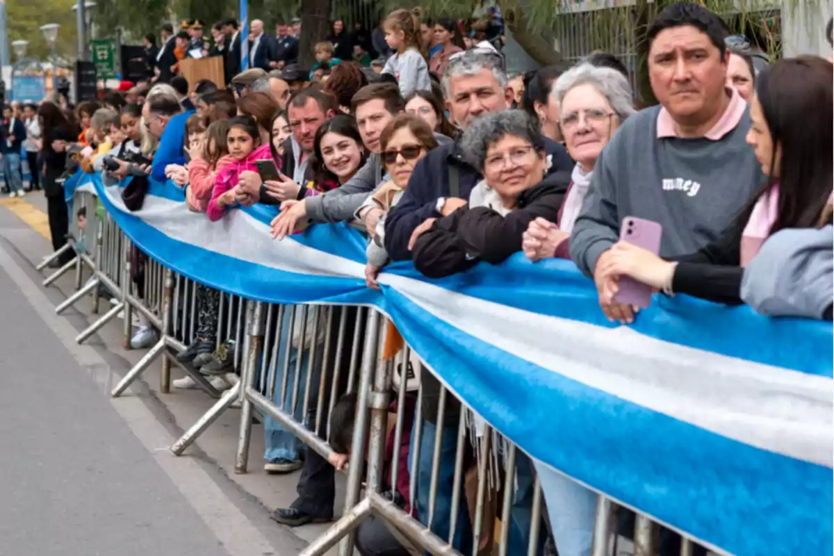 People of different ages watch a parade from behind a fence decorated with light blue and white fabric. People of different ages watch a parade from behind a fence decorated with light blue and white fabric.