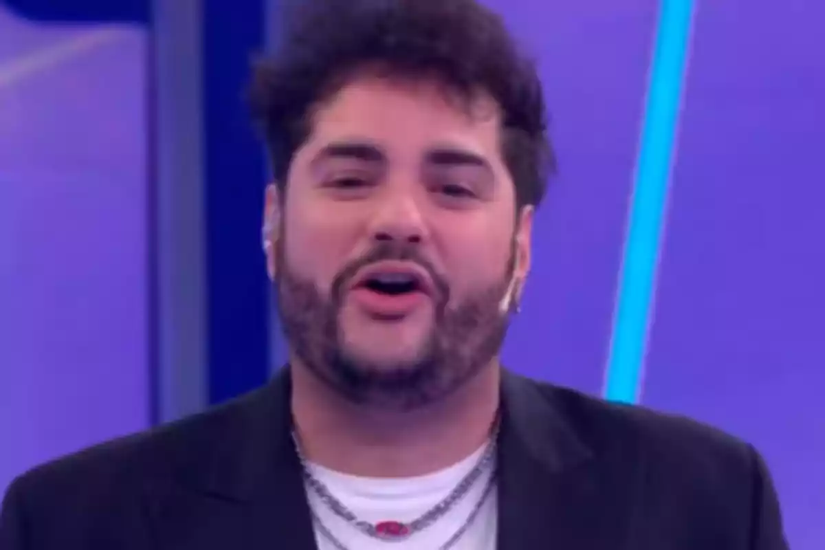 Hombre con barba y cabello rizado usando chaqueta negra y camiseta blanca en un set de televisión con fondo morado Hombre con barba y cabello rizado usando chaqueta negra y camiseta blanca en un set de televisión con fondo morado
