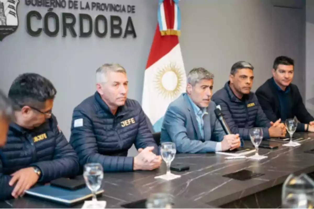 Five men seated at a table during a press conference in an official government room in Córdoba with the Argentine flag in the background