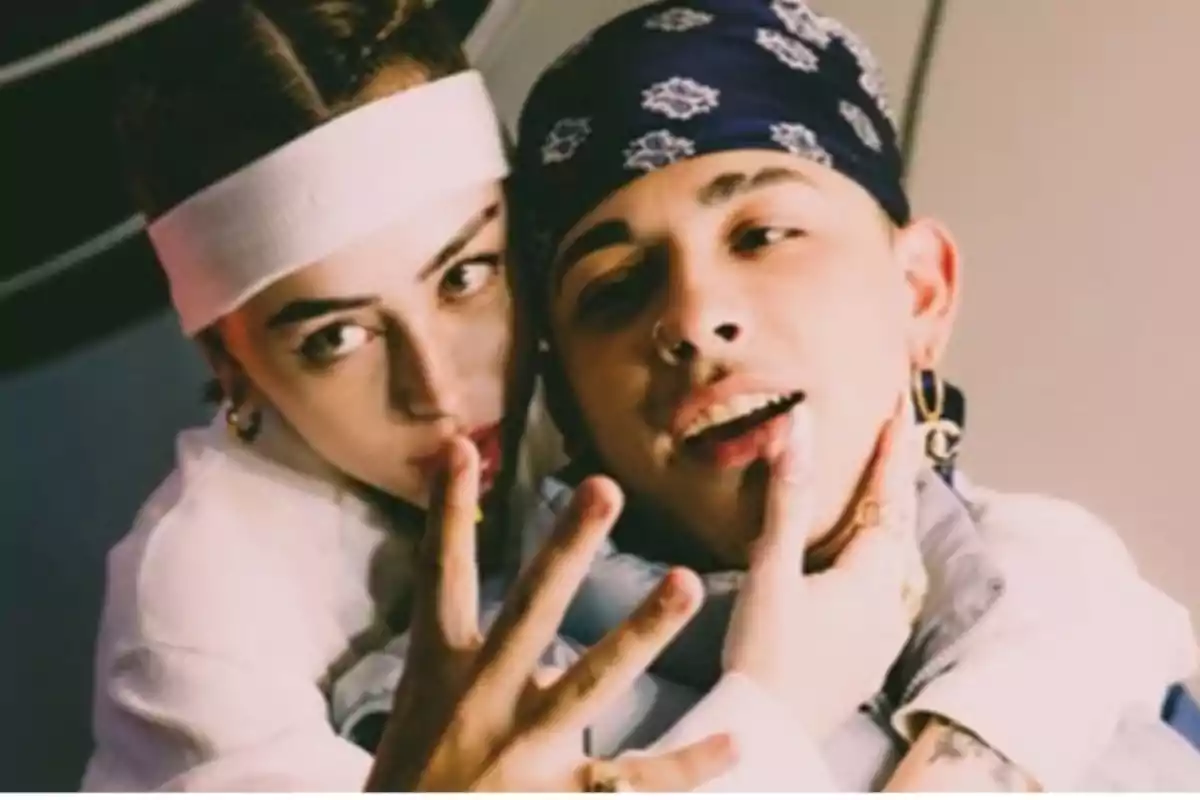 Two people pose together looking at the camera, one wearing a white headband and the other a dark patterned scarf, both with relaxed expressions and dressed in casual clothing.