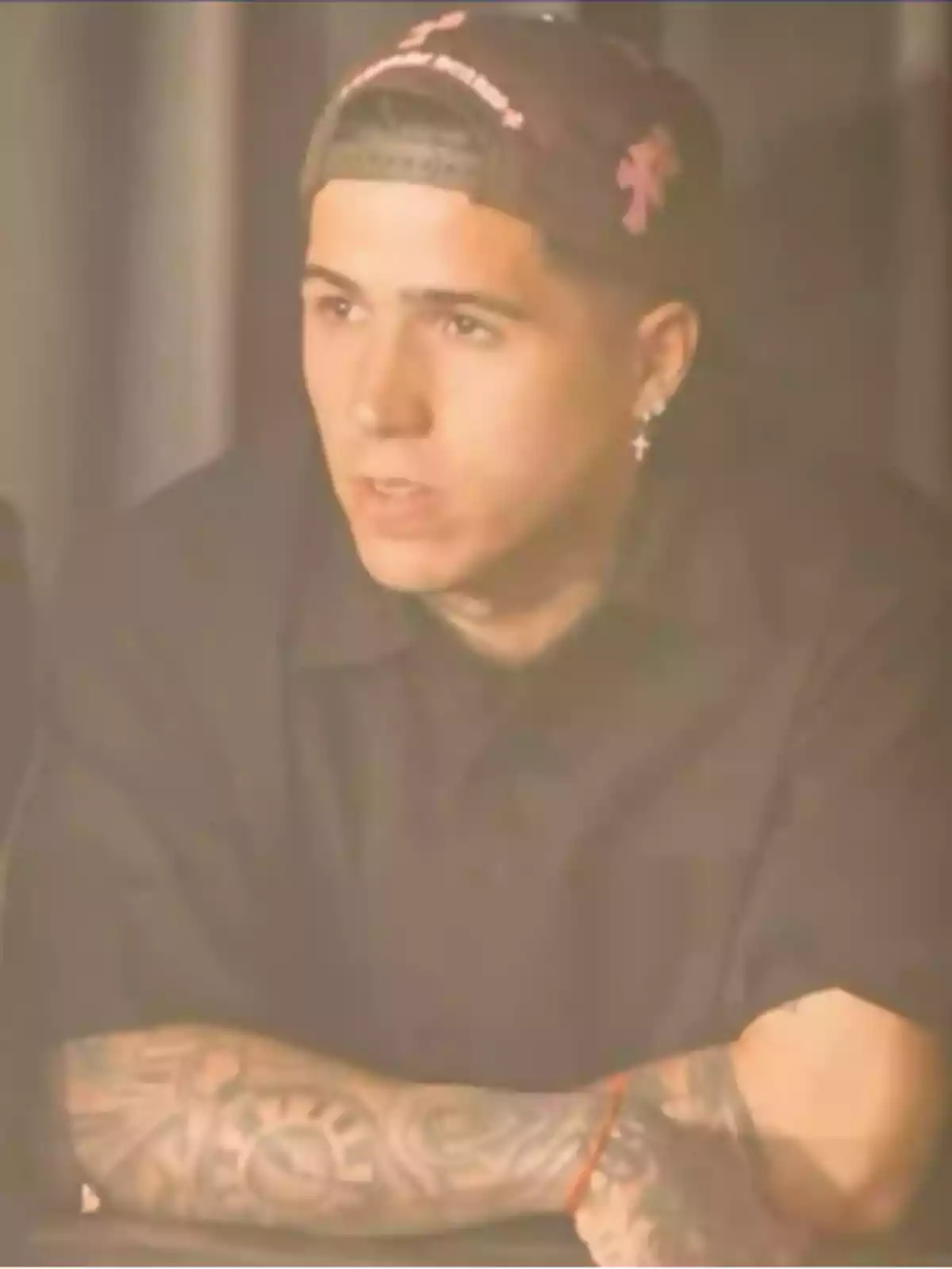 Young man with a backwards cap, dark t-jersey, tattoos on his arm, and a cross earring, sitting with his arms crossed Young man with a backwards cap, dark t-jersey, tattoos on his arm, and a cross earring, sitting with his arms crossed