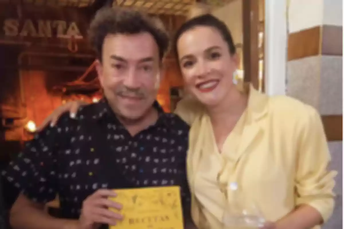 Two people smile and pose together indoors, one of them holding a yellow book. Two people smile and pose together indoors, one of them holding a yellow book.