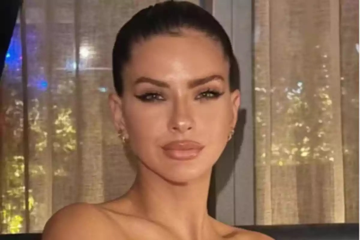 Mujer de cabello oscuro recogido con maquillaje elegante posando frente a una cortina beige