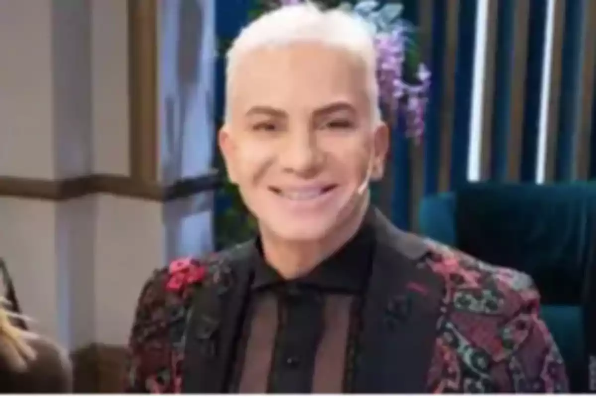 Persona de cabello corto y canoso sonríe mientras usa un saco colorido con detalles florales y una camisa negra en un ambiente interior elegante