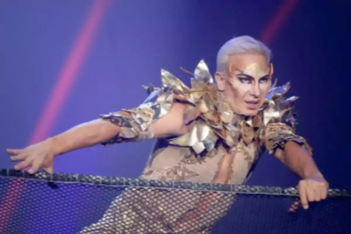 Persona con cabello platinado y maquillaje artístico usando un vestuario dorado con detalles metálicos en un escenario iluminado Persona con cabello platinado y maquillaje artístico usando un vestuario dorado con detalles metálicos en un escenario iluminado