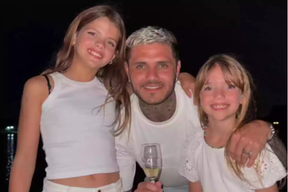 Tres personas sonríen abrazadas, dos niñas y un hombre con una copa en la mano, todos vestidos de blanco y con fondo oscuro Tres personas sonríen abrazadas, dos niñas y un hombre con una copa en la mano, todos vestidos de blanco y con fondo oscuro