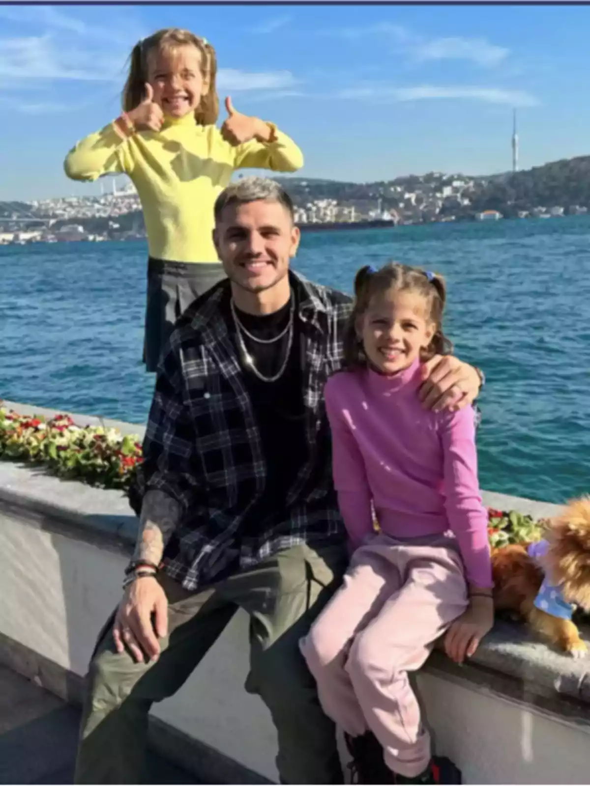 Un hombre y dos niñas sonríen junto a un perro pequeño frente a un paisaje de mar y ciudad en un día soleado Un hombre y dos niñas sonríen junto a un perro pequeño frente a un paisaje de mar y ciudad en un día soleado