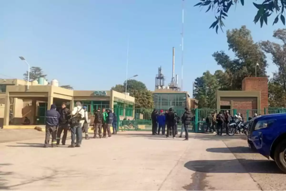 Several people gathered in front of the entrance to an industrial plant with green gates and parked vehicles. Several people gathered in front of the entrance to an industrial plant with green gates and parked vehicles.