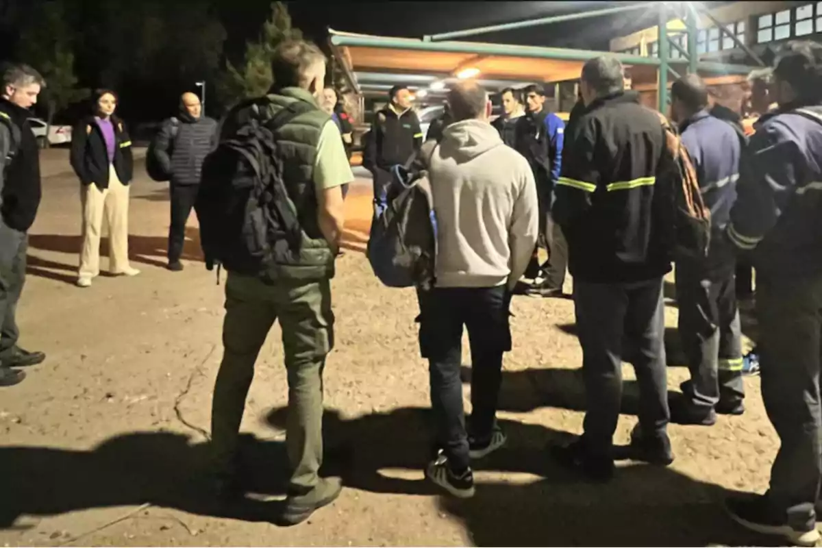 A group of people gathered outdoors at night under a lit structure, some wearing backpacks and casual clothing. A group of people gathered outdoors at night under a lit structure, some wearing backpacks and casual clothing.