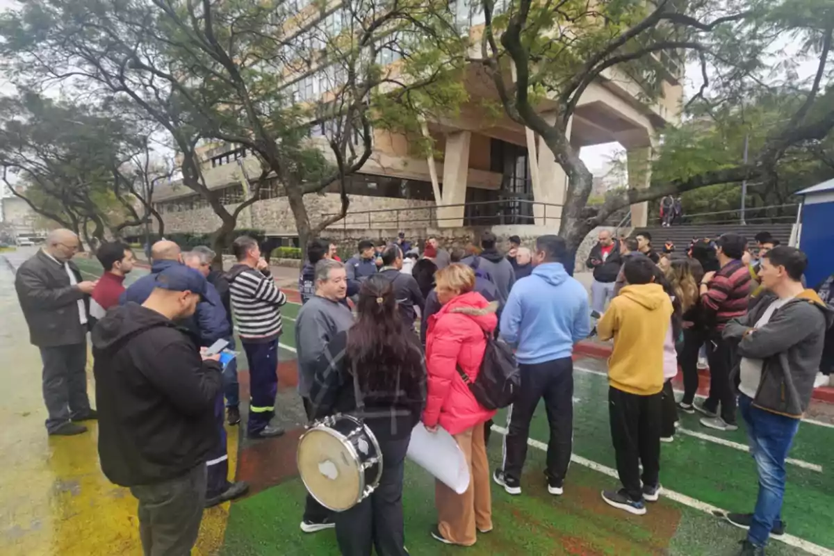 A group of people gathered outdoors in front of a building surrounded by trees, some talking and others observing, one person holding a drum.
