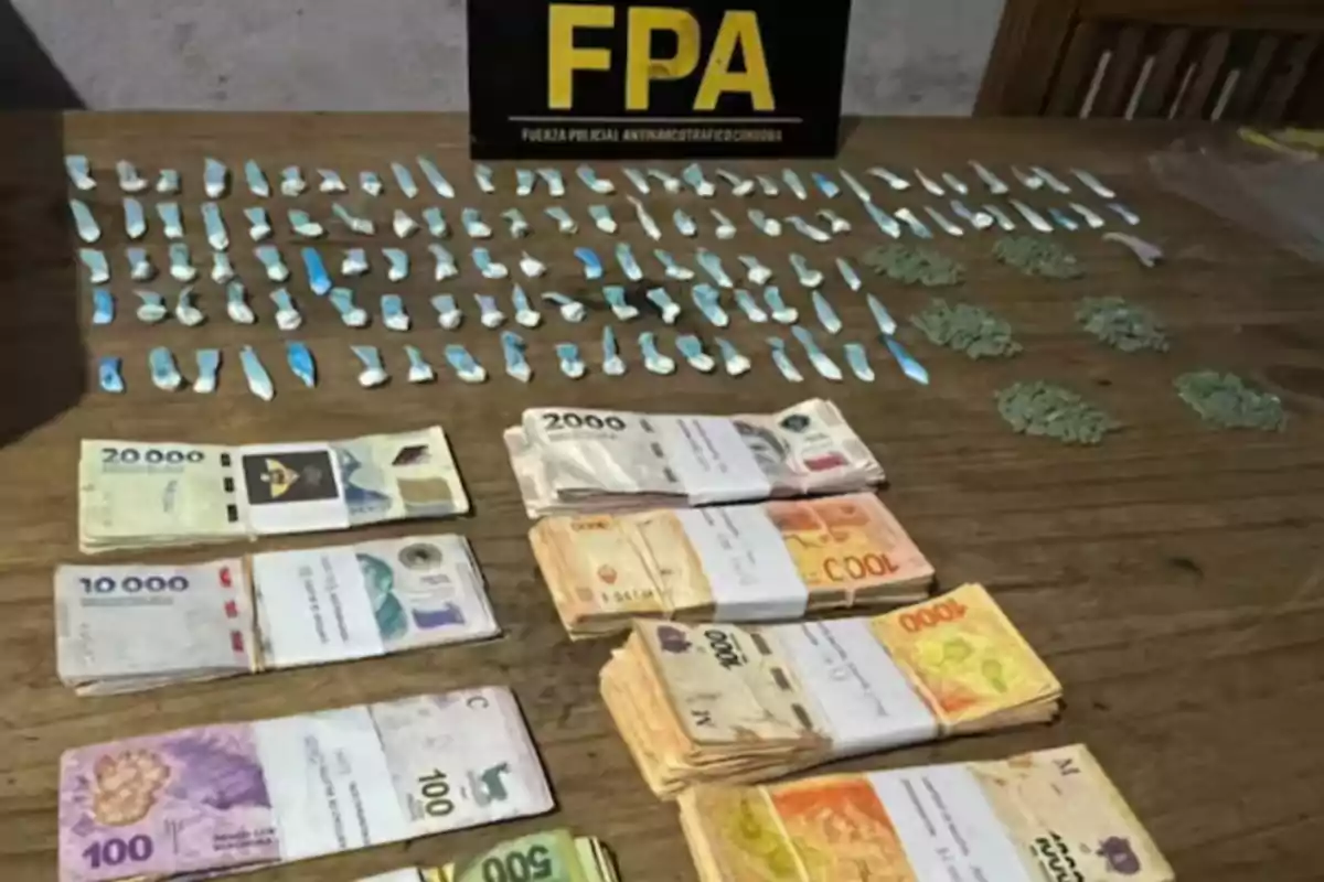 On a wooden table, there are several bundles of bills of different denominations, small bags with substances and pills organized in rows, next to a black and yellow sign with the initials FPA. On a wooden table, there are several bundles of bills of different denominations, small bags with substances and pills organized in rows, next to a black and yellow sign with the initials FPA.