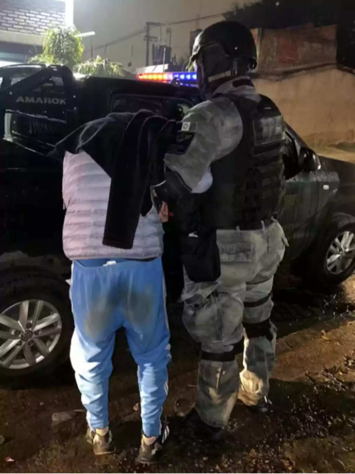 A police officer in tactical uniform detains a person wearing blue pants and a light jacket next to a black pickup truck with its lights on on a street at night. A police officer in tactical uniform detains a person wearing blue pants and a light jacket next to a black pickup truck with its lights on on a street at night.