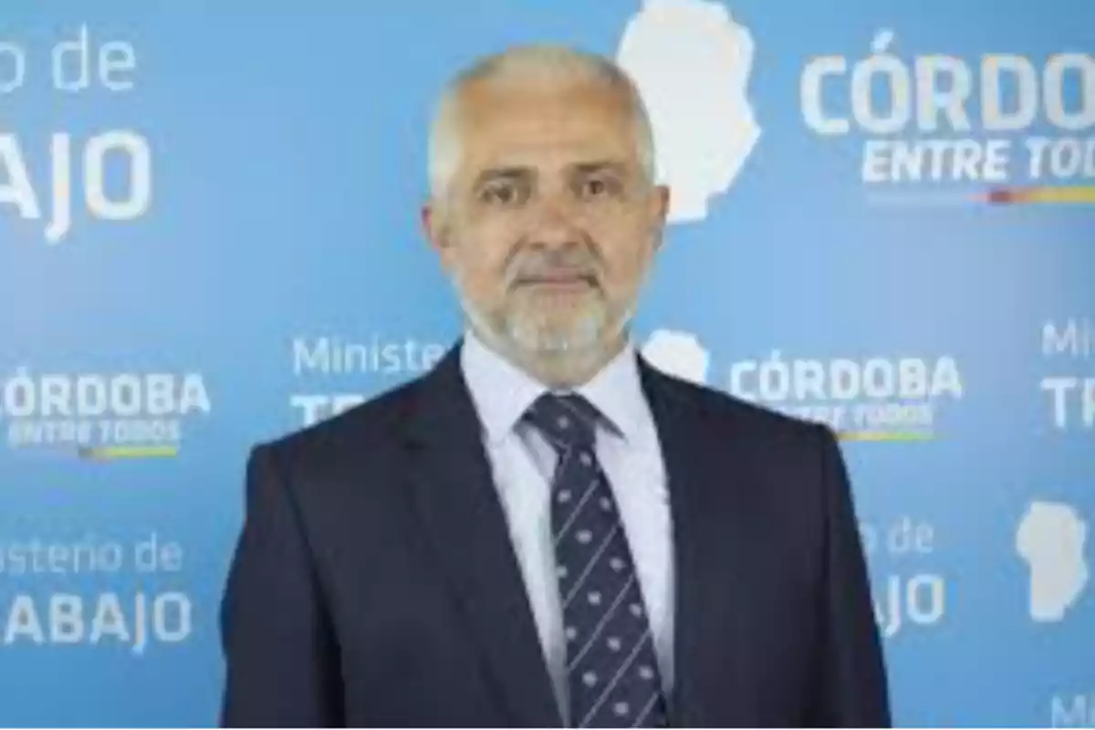 Hombre de cabello canoso y barba, vestido con traje y corbata, posando frente a un fondo azul con textos relacionados al Ministerio de Trabajo de Córdoba Hombre de cabello canoso y barba, vestido con traje y corbata, posando frente a un fondo azul con textos relacionados al Ministerio de Trabajo de Córdoba