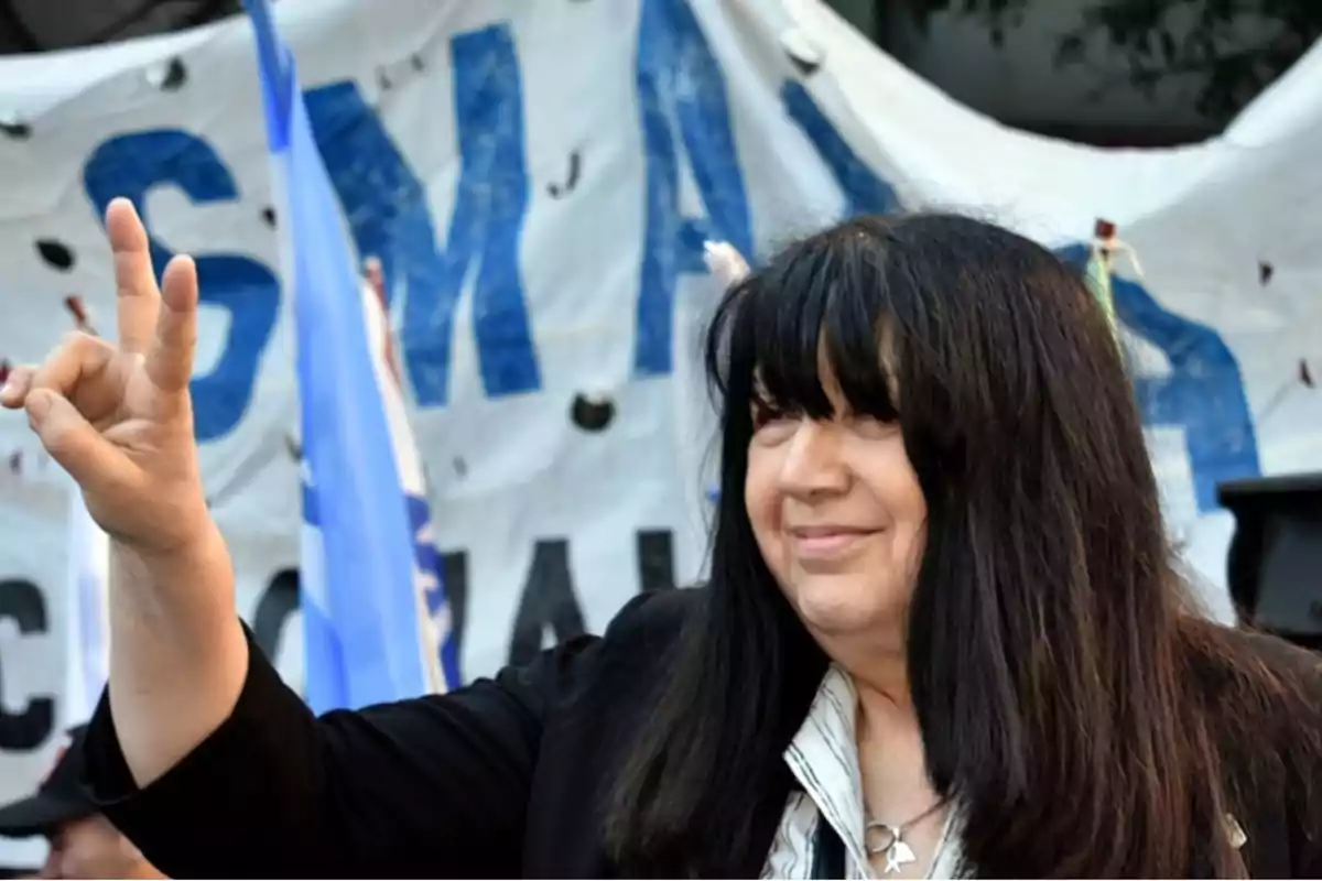 Long-haired, dark-haired woman smiles while waving at an outdoor event with a flag and a sign in the background. Long-haired, dark-haired woman smiles while waving at an outdoor event with a flag and a sign in the background.