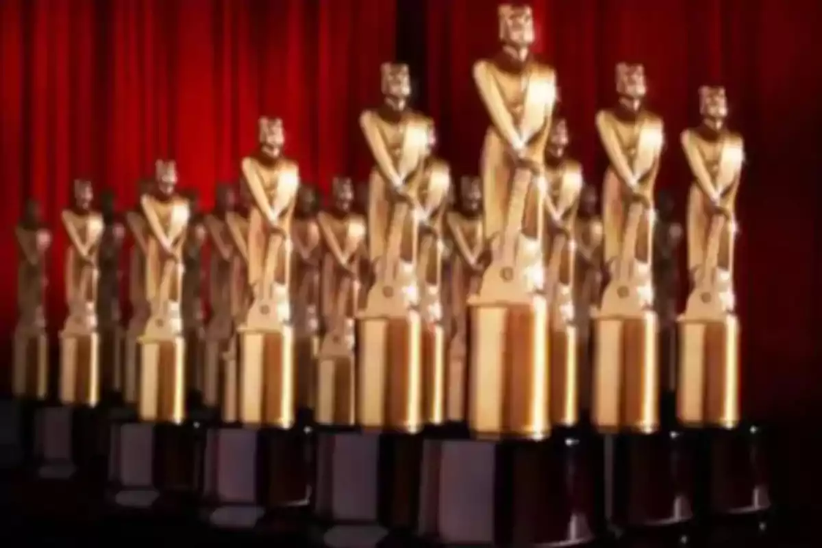 Several golden trophies in the shape of human figures with a background of red curtains