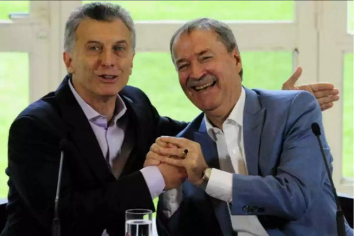 Two men in suits smile and greet each other enthusiastically while sitting in front of microphones and a glass of water. Two men in suits smile and greet each other enthusiastically while sitting in front of microphones and a glass of water.