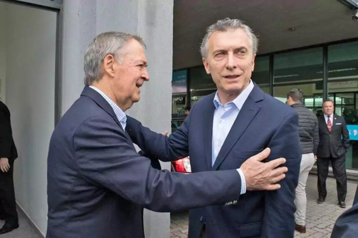 Two men in suits greet each other cordially at the entrance of a building while other people watch in the background. Two men in suits greet each other cordially at the entrance of a building while other people watch in the background.